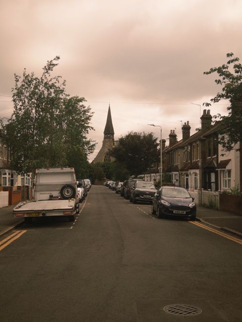 A residential street scene during daytime with a row of parked cars on either side of the narrow road, including a white van with its rear hatch open, revealing packing and moving supplies such as car